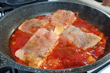 Salmon fish pieces stewed in a fresh tomato, olive oil with lemon, garlic, sweet bell pepper and rosemary.  Dish is in black frying pan on the white gas stove. Mediterranean spicy fish dish 