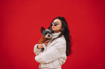 Attractive girl in casual clothes isolated on red background with a dog in her arms, looking aside. Pets concept.