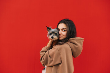 Portrait of a cute woman with a small dog in her arms isolated on a red background, looking at the camera and smiling. Copy space