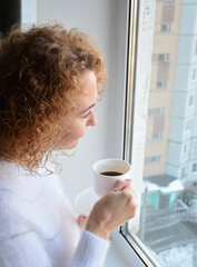 a girl with curly hair in a white sweater drinks coffee from a white cup and looks thoughtfully out the window.