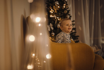 Happy child sits on an armchair with decoration and Christmas tree in the background.