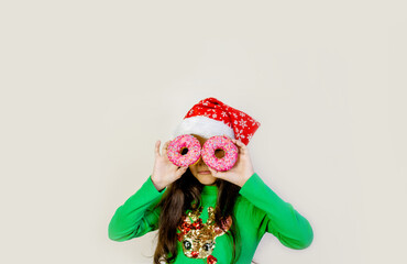 Cute dak hair girl in santa hat smiling, having fun and looking through two donuts on her eyes. Sweets. Grey studio background.