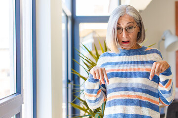 middle age woman feeling shocked, open-mouthed and amazed, looking and pointing downwards in...