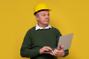 Senior foreman with special hard hat working using laptop. Studio shot on yellow wall.