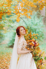 Obraz na płótnie Canvas Sweet red-haired girl in a wedding dress and with a bouquet in her hands posing on the background of the autumn garden.