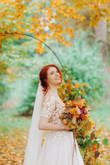 Cute red-haired girl in a wedding dress and with a bouquet in her hands posing on the background of the autumn garden.