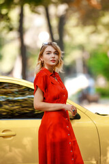 Young attractive and dreamy business woman in red dress near a yellow car in a city park