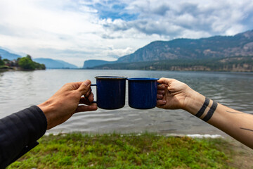 Selective focus of two different people clinching their cups of coffee, or tea to each other with a nature background with a lake and mountains