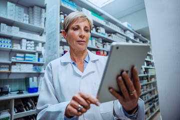 Naklejka na ściany i meble Caucasian senior pharmacist checking medication prescriptions on digital tablet standing in pharmacy 