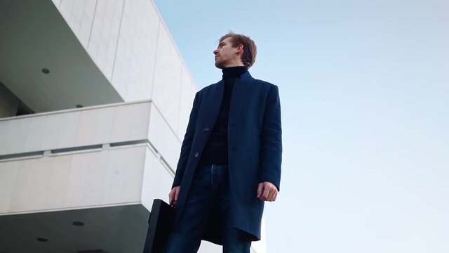 Young confident business man stands in downtown. A successful entrepreneur with a folder of documents in his hands near the office building. Motivation. Bottom view. Slow motion