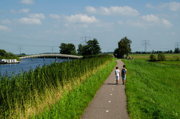 Obraz premium Kinderdijk, The Netherlands, August 2019. A small road winds through the Dutch countryside, lined with green grass and a canal, some boys are walking along it. On the left a white bridge.