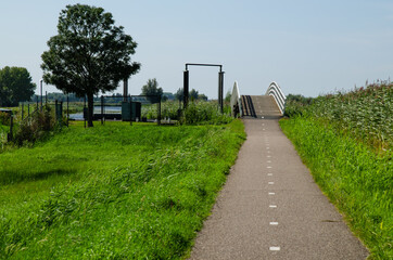 Kinderdijk, The Netherlands, August 2019. A small road winds through the Dutch countryside, lined with green grass and a canal, in the background a bridge with a white parapet.