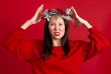 close-up portrait of a nervous, aggressive Asian young woman in a Christmas wreath with hands holding her head, on a red background.