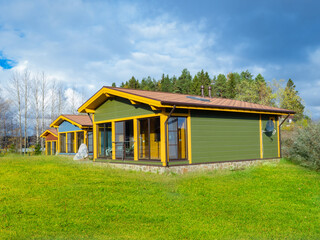 Country cottages in a minimalistic style. Wooden cottages on a blue sky background. Forest behind cottage community. Several one-story houses stand side by side. Lawn in front of village houses.