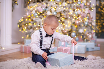 Christmas tree, a boy with Down syndrome sits on the floor and holds a box with a gift in his...