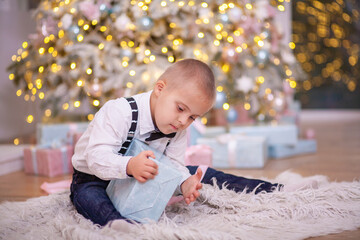 Christmas tree, a boy with Down syndrome sits on the floor and holds a box with a gift in his...
