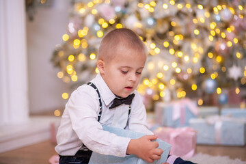 Christmas tree, a boy with Down syndrome sits on the floor and holds a box with a gift in his...