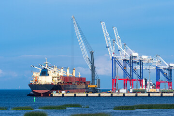 Naklejka premium Cargo seaport. Terminal for loading sea containers. Loading cranes on shore of cargo port. Crane is loading something onto ship. Marine ship during shipment. Summer day. Unloading a sea vessel