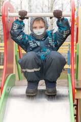 Preschooler boy alone on the playground wearing a mask and autumn clothes. Concept - children in quarantine, coronavirus pandemic, virus protection, isolation and social distance with covid 19.