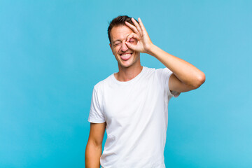 young handsome man smiling happily with funny face, joking and looking through peephole, spying on...