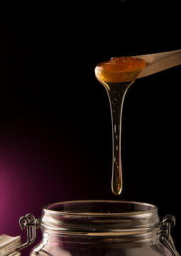 Product Shot Of A Spoon With Honey Dripping Into A Jar, Isolated In A Black Background With A Purple Backlight