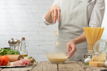 Chef in white uniform pours cheese in sauce into glass bowl for cooking pasta alla carbonara. Backstage of preparing traditional italian dish on white background. Frozen motion. Recipe book. Close-up.