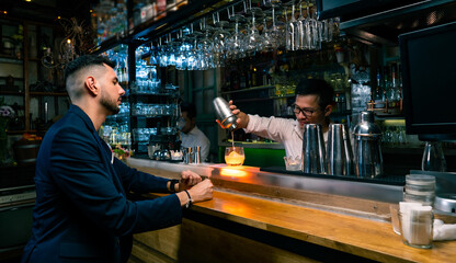 Portrait of Handsome Caucasian man sitting at bar counter and waiting tasty cocktail from barman in nightclub. Asian male mixologist bartender preparing mixed alcohol drink on rocks glass for customer