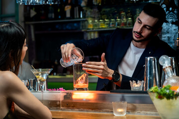Professional male mixologist bartender preparing mixed alcoholic cocktail drink serving to woman sitting at bar counter in nightclub. Caucasian barman pouring mixed liquor into decorative rocks glass.