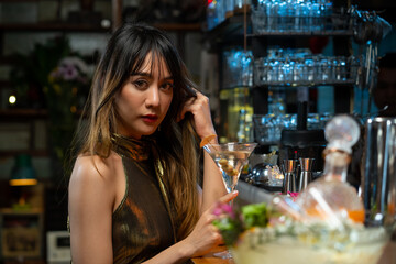 Portrait of Beautiful Asian woman standing at bar counter holding martini glass and enjoy drinking alcoholic cocktail drink from barman in nightclub. Nightlife party celebration and holiday concept