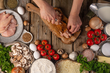 Chef hands cuts chicken fillet on wooden chopped board on table with variety of ingredients background. Concept of cooking process. Backstage of preparing tasty meal. View from above. Flat lay.