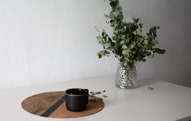 Taza de cerámica negra junto a un ramo de hojas de eucaliptus secas sobre una mesa blanca moderna...