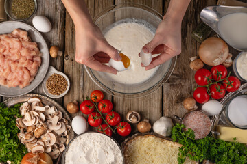 Chef hands breaks egg into glass bowl with milk on wooden table with variety of ingredients background. Concept of cooking process. Backstage of preparing tasty meal. View from above. Frozen motion.