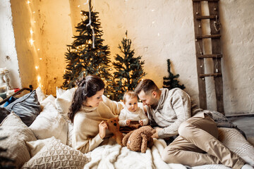 Happy parents with little daughter lay at the bed near decorated Christmas tree, smiling, enjoy...