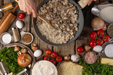 Chef hands mix sour cream with chicken in pan on wooden table with variety of ingredients background. Concept of cooking process. Backstage of preparing tasty meal. View from above. Frozen motion.