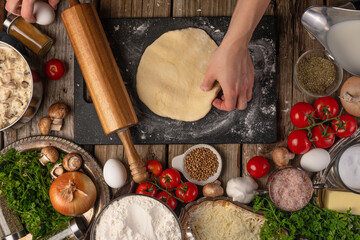 Chef hands with the dough and rolling pin on wooden table with variety of ingredients background. Concept of cooking process. Backstage of preparing tasty meal. View from above.