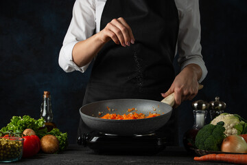 Professional chef salts frying carrot in the pan on variety of ingredients background. Backstage of preparing appetizing food. Concept of cooking process. Frozen motion.