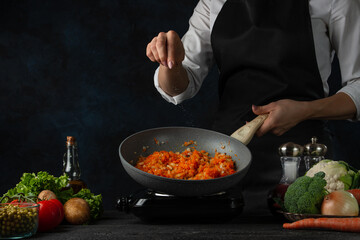 Close-up view of professional chef salts frying carrot in the pan on variety of ingredients background. Backstage of preparing appetizing food. Concept of cooking process. Frozen motion.