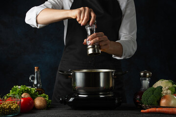Professional chef pours spices into boiling water in the pot on variety of ingredients background. Backstage of preparing appetizing food. Concept of cooking process. Frozen motion.