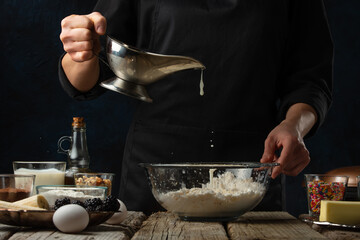 Pastry chef pours milk into glass bowl with flour on table with ingredients for cooking on dark blue background. Backstage of preparing sweet waffles. Frozen motion. Dessert from recipe book.