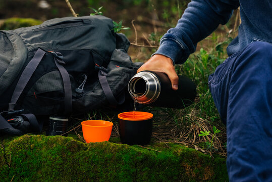 Drinking tea during hike. Man hand pouring hot tea from black thermos bottle into 2 cup's in the woods. Refreshment during hiking. Camping equipment