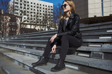 Woman in leather jacket sits on the stairs near the building in the autumn on the street