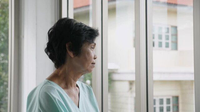 Indoors, close up, side view portrait of worried senior Asian woman wear green patient outfit, stand near the windows, look outside sadly