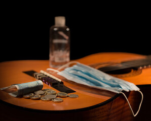 Acoustic Guitar, Face Mask, Hand Sanitiser and Cash on a Black Background.
Concept for lack or earnings, cancelled shows, gigs, tours for performing musicians. Or requirements for live music events.