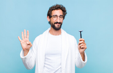 young bearded man smiling and looking friendly, showing number four or fourth with hand forward,...