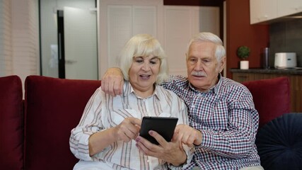 Happy smiling senior couple with tablet pc computer at home. Cheerful retired husband and wife hugging and laughing using digital tablet resting on sofa in cozy living room. 6k downscale, slow motion - Powered by Adobe
