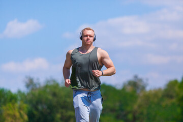 Front view of muscular young man in headphones runs in a summer Park. Running in the fresh air
