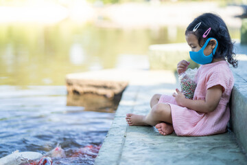 Cute little girl sitting at pond wearing protective face mask feeding carp fishes