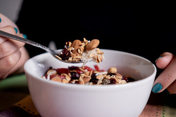 Person eating homemade oatmeal with banana, strawberries, granola, blueberry, coconut and almonds with a spoon inside a white plate.