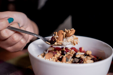Person eating homemade oatmeal with banana, strawberries, granola, blueberry, coconut and almonds with a spoon inside a white plate.