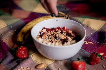 Exquisite oatmeal with banana, strawberries with one hand pouring granola, blueberry, coconut and almonds on top, inside a white plate.